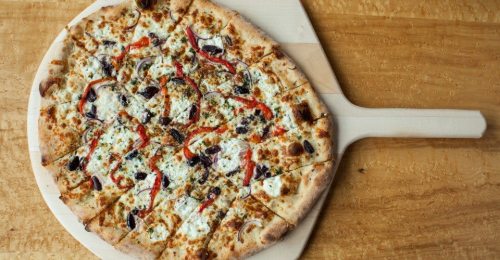 A freshly baked pizza topped with cheese, red peppers, onions, and olives on a wooden pizza peel.