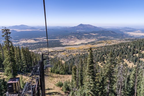 A scenic view from a ski lift, overlooking a valley filled with trees and distant mountains under a clear blue sky.