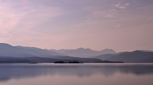 Serene lake scene with soft purple hues, distant mountains, and calm water reflecting the sky.