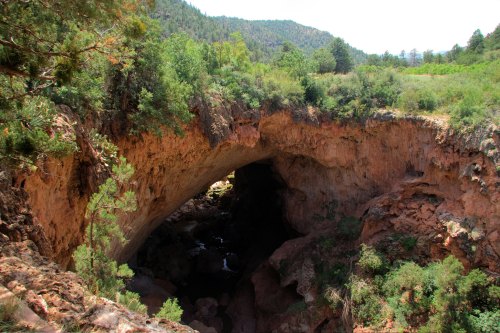 Natural stone arch bridge surrounded by lush greenery and rocky terrain, with a stream flowing below.