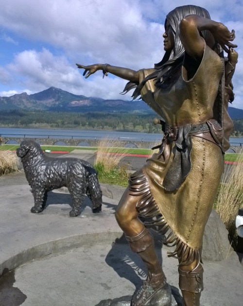 A bronze statue of a woman in traditional attire pointing, with a dog statue beside her and mountains in the background.