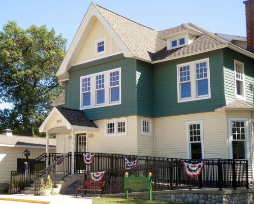 A two-story house with green and yellow siding, featuring a porch and decorative bunting, surrounded by trees.