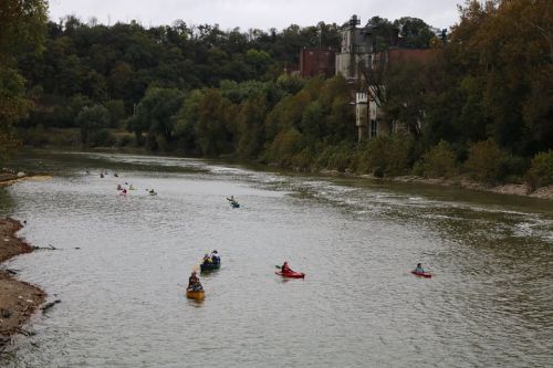 A group of kayakers paddles along a calm river surrounded by trees and a distant industrial building.