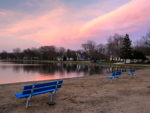 Three blue benches on a sandy beach by a calm lake, with a colorful sunset and trees in the background.