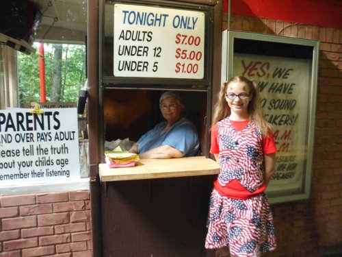 A girl in a patriotic outfit stands next to a ticket booth with a smiling woman inside, surrounded by signs.