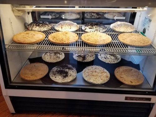 A display case filled with various types of pies, including cream and crumb crusts, arranged on wire shelves.