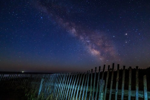A starry night sky with the Milky Way above a wooden fence near a calm body of water.