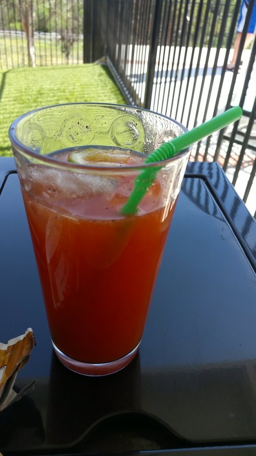 A refreshing drink with a green straw, served in a clear glass, sits on a black table by a poolside.