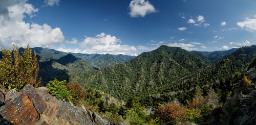 A panoramic view of lush green mountains under a blue sky with scattered clouds. Rocky foreground adds depth to the scene.