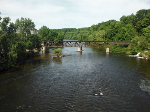 A tranquil river flows under a metal bridge, surrounded by lush green trees and a clear blue sky.