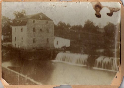 A vintage photo of a stone mill beside a waterfall, with trees in the background and a slightly faded appearance.