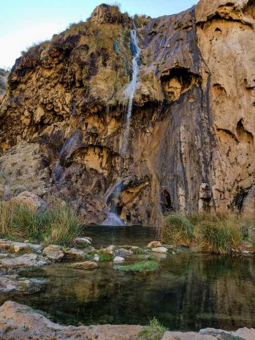 A serene waterfall cascading down a rocky cliff into a clear pool surrounded by grass and stones.