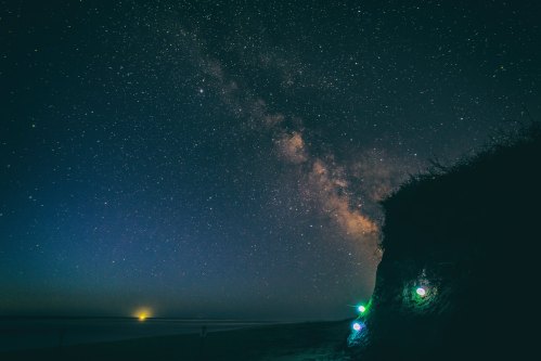 A starry night sky with the Milky Way over a beach, featuring glowing lights on a grassy dune.