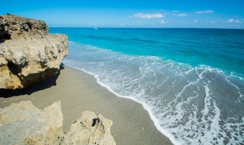 A serene beach scene with gentle waves lapping at the shore, rocky formations, and a clear blue sky.