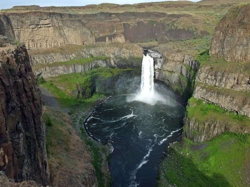 Aerial view of a waterfall cascading into a circular pool, surrounded by rocky cliffs and lush greenery.