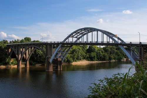 A steel arch bridge spans a river, surrounded by lush greenery and a clear blue sky.