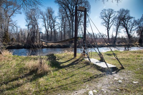 A rustic suspension bridge over a river, surrounded by tall trees and grassy banks under a clear blue sky.