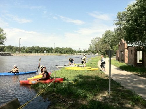 People kayaking along a river, with some preparing to launch from the shore on a sunny day.