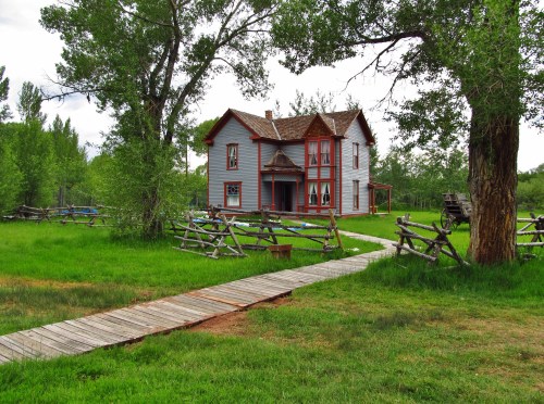 A charming two-story house with red trim, surrounded by green grass and trees, with a wooden walkway leading to it.