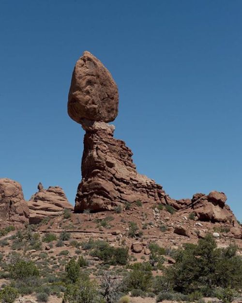 A large, balanced rock perched atop a tall stone formation against a clear blue sky in a desert landscape.