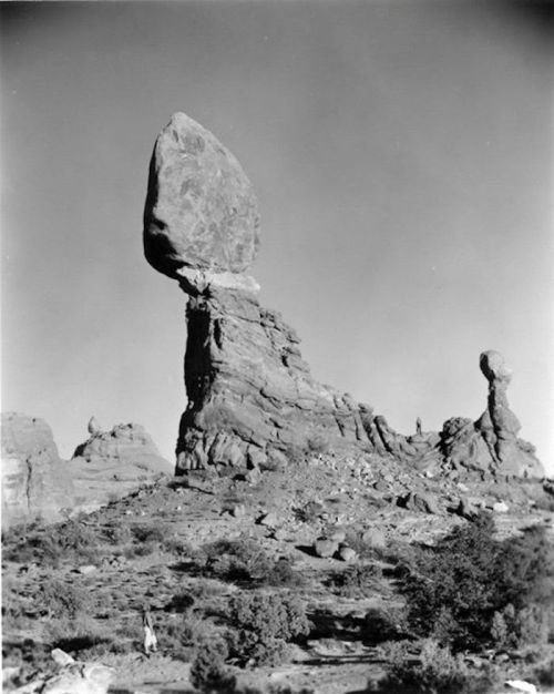 A large, balanced rock perched atop a tall stone formation in a desert landscape, with smaller rocks in the background.