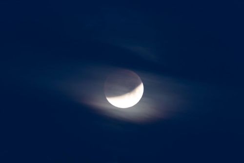 A partially eclipsed moon partially obscured by dark clouds against a deep blue sky.