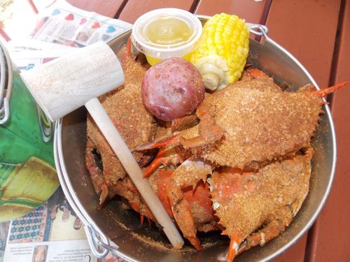 A bucket filled with seasoned crabs, corn on the cob, a potato, and a small container of sauce on a wooden table.