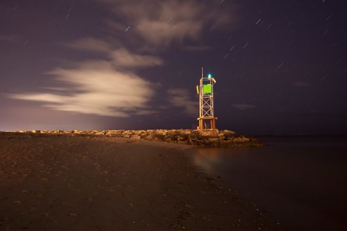 A lighthouse on a rocky shore at night, with stars and clouds in the sky, reflecting in the calm water.