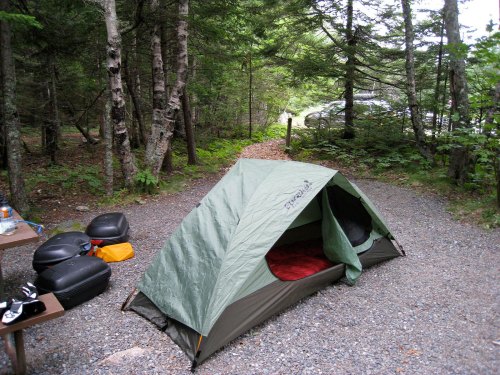 A green tent set up in a wooded campsite, with a gravel path and some camping gear nearby.