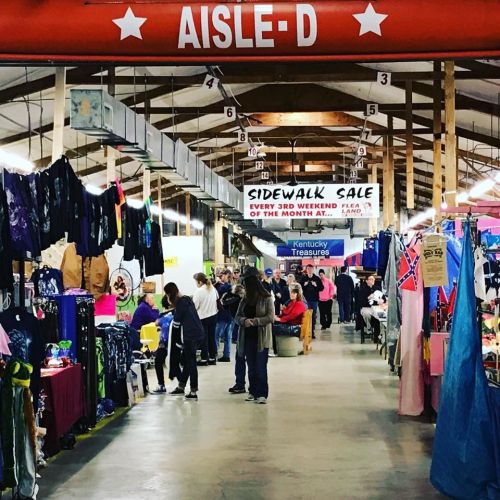 A bustling indoor market aisle with vendors, shoppers, and colorful merchandise under a sign for a sidewalk sale.