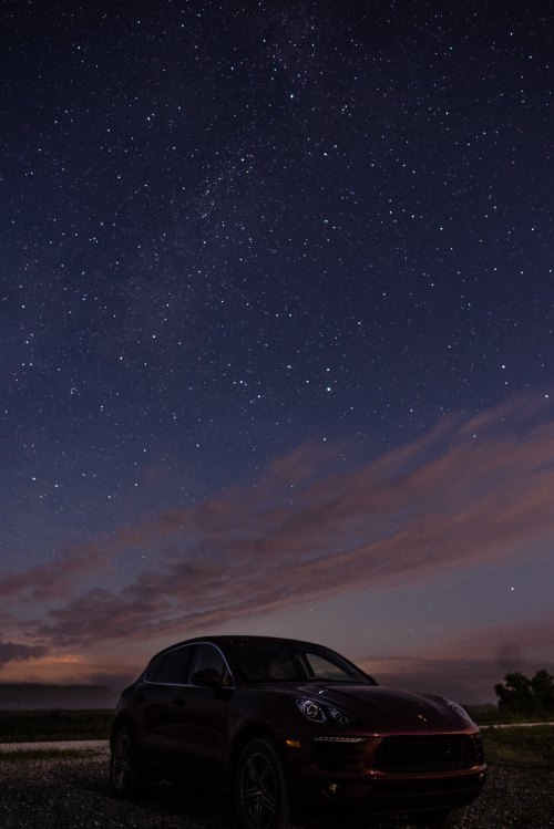 A car parked under a starry night sky with the Milky Way visible and clouds in the background.