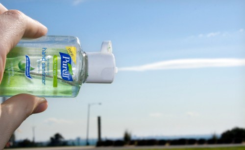 A hand holding a bottle of Purell hand sanitizer against a clear blue sky with wispy clouds.