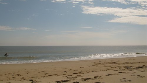 A serene beach scene with gentle waves, a clear sky, and a lone surfer in the water.