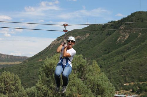 A person in a helmet zip-lining over a green landscape with mountains in the background, smiling and enjoying the ride.