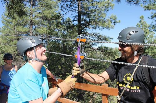 Two people in helmets smile while connecting gear on a zip line, surrounded by trees and a sunny sky.