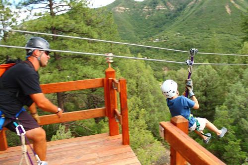 A person zip-lining from a platform while another person assists on the side, surrounded by green mountains.
