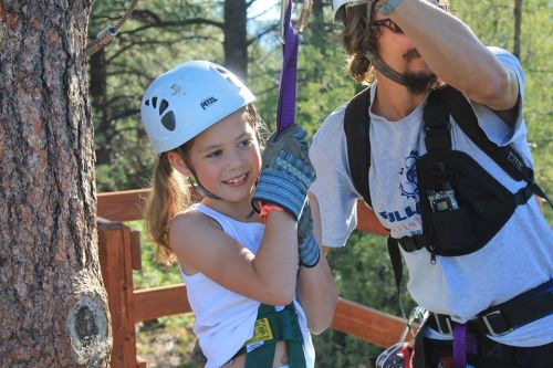 A young girl in a helmet smiles while holding onto a rope, with an adult assisting her in a forested area.