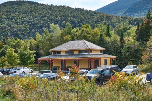 A yellow building with a porch surrounded by trees and mountains, with parked cars in the foreground.
