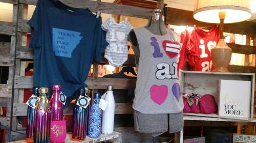 Display of clothing and accessories, including shirts, a onesie, and water bottles, in a rustic shop setting.
