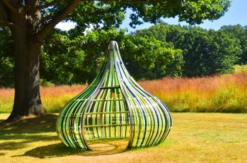 A colorful, teardrop-shaped sculpture made of wooden slats, set in a grassy field under a large tree.