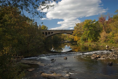 A stone bridge arches over a calm river, surrounded by colorful autumn trees and a clear blue sky.
