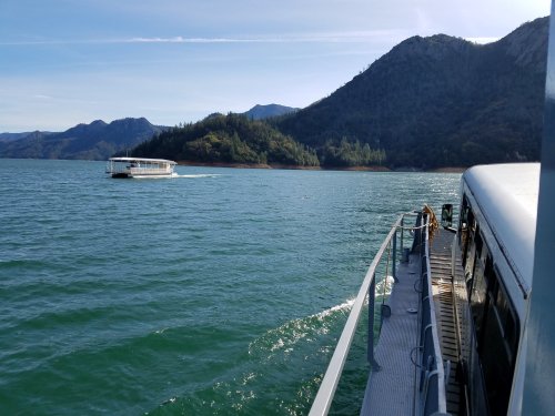 A boat glides across a serene lake, surrounded by mountains and lush greenery under a clear blue sky.