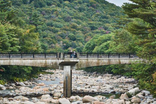 A bridge spans a rocky riverbed, surrounded by lush green trees and a hilly landscape in the background.