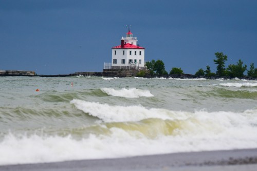 A white lighthouse with a red roof stands on a rocky shore, with waves crashing in the foreground under a cloudy sky.