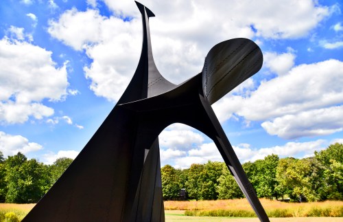A large, abstract black sculpture against a blue sky with fluffy clouds and green trees in the background.