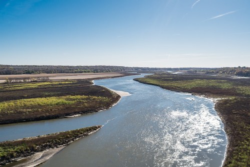 A winding river flows through a lush landscape under a clear blue sky, surrounded by greenery and open fields.