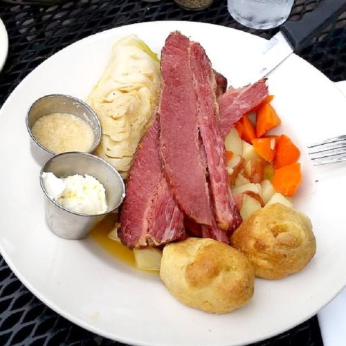 A plate of corned beef with cabbage, carrots, potatoes, and two small rolls, served with mustard and a creamy dip.