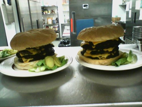 Two large burgers stacked high with multiple patties and cheese, served with pickles and lettuce on the side.