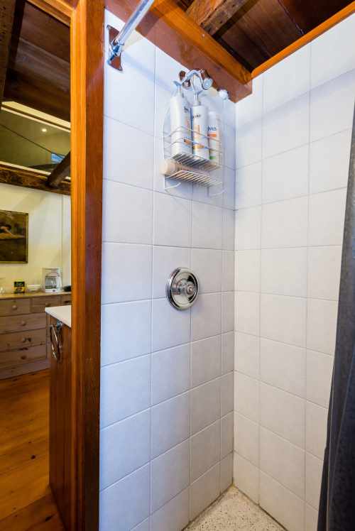 A clean shower with white tiles, a showerhead, and a shelf holding shampoo bottles. Wooden beams are visible above.
