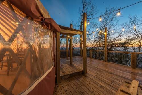 A wooden deck with string lights at sunset, featuring a cozy yurt structure and trees in the background.